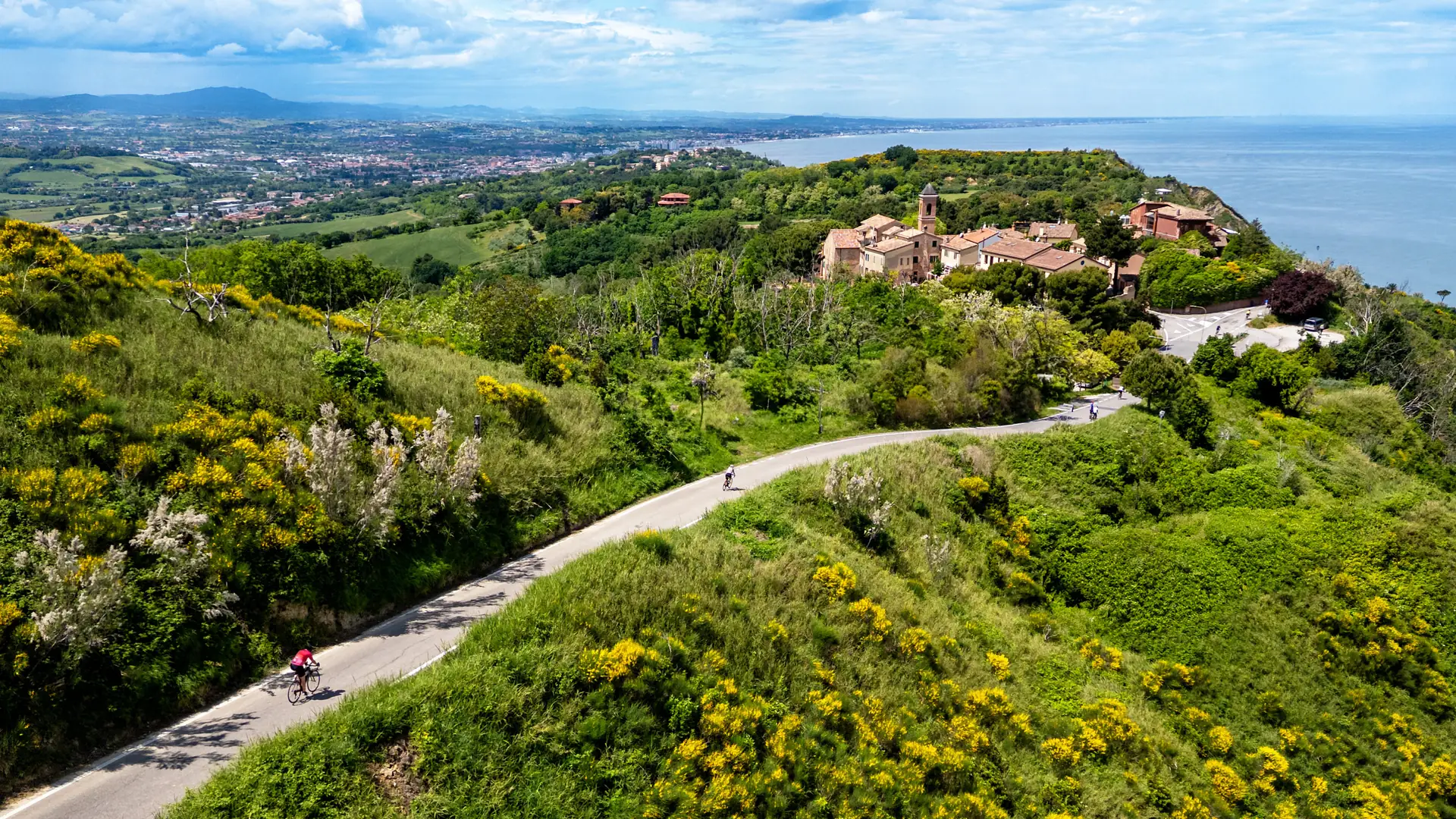 Ciclisti su una strada panoramica tra le ginestre, con un borgo sulla collina e il mare sullo sfondo